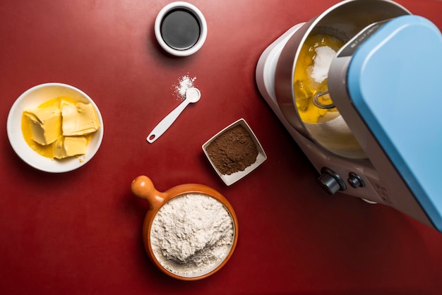 Top view of an electric mixer flour butter cocoa and other ingredients on a red table