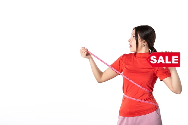 Top view of emotional shocked beautiful girl in redorange blouse holding bank card measuring herself with metre holding sale sign on white background