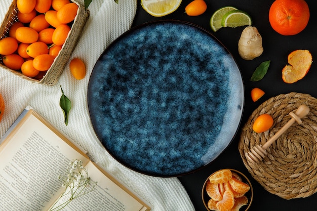 Top view of empty plate with tangerine slices and open book with basket of kumquat and little flower on book and lemon half lime slices orange ginger with trivet honey dipper on cloth