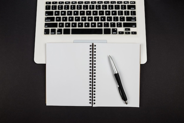 Top view of empty white notepad with pen and laptop on black background copy space