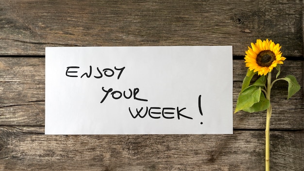 Photo top view of enjoy your week message written on white paper card laying on a textured wooden desk next to a beautiful blooming sunflower.