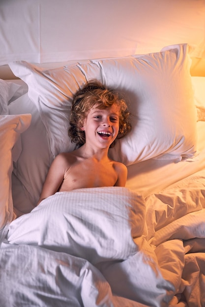 Top view of excited shirtless boy with curly hair lying under warm blanket on soft bed and laughing in dim bedroom at home