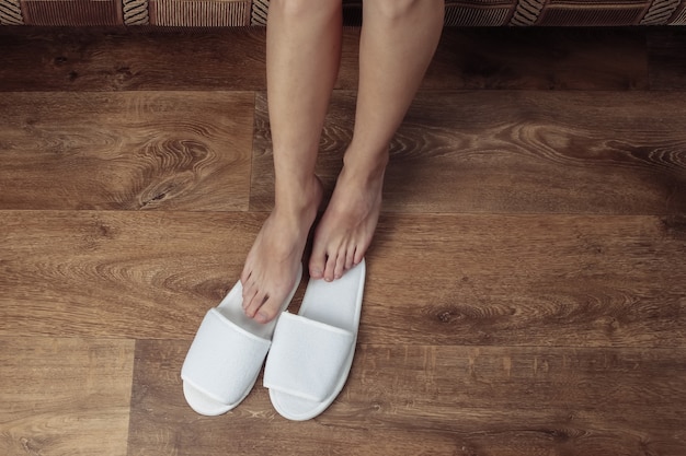 Top view of female bare legs with white slippers on a wooden floor.