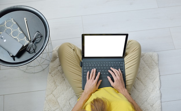 Top view of female freelancer sitting on floor near sofa at home and working with blank laptop screen