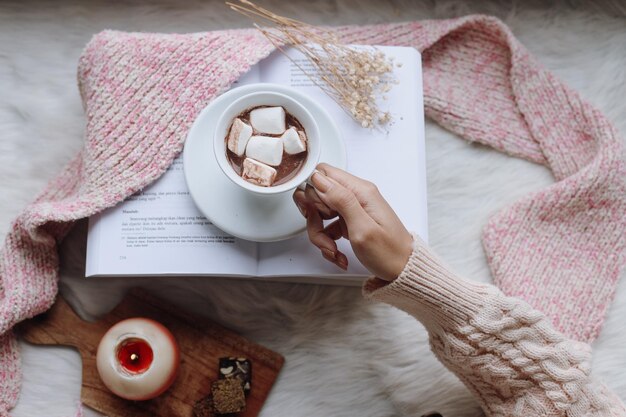 Top view female hand holding cup of hot chocolate marshmallow on fur rug with pink scarf