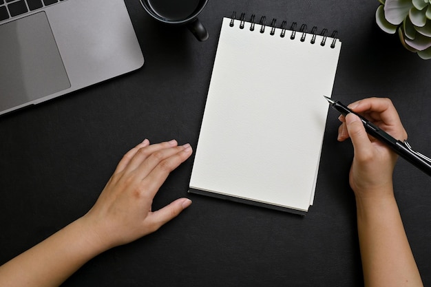 Top view A female hand holding pen taking notes on spiral notepad in her modern black desk