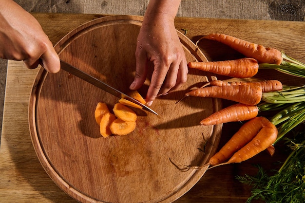 Top view of female hands cutting, chopping, slicing fresh raw baby carrot on a wooden board. Close-up of hand with knife cutting fresh vegetable. Flat lay food background