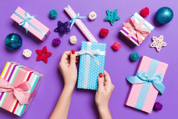 Top view of female hands holding a Christmas present on festive purple background. Holiday decorations, toys and balls. New Year holiday concept.