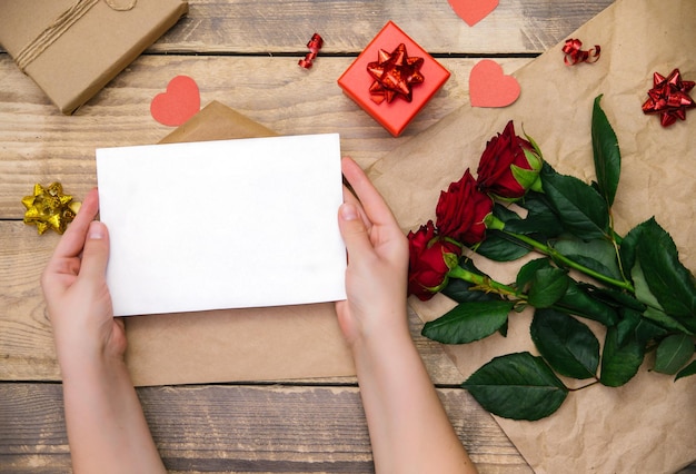 Top view, female hands opening an envelope with an invitation or a postcard.Red roses, gift box on a wooden background.