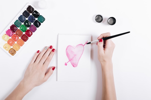 Top view of a female hands with red manicure painted watercolor heart in notebook