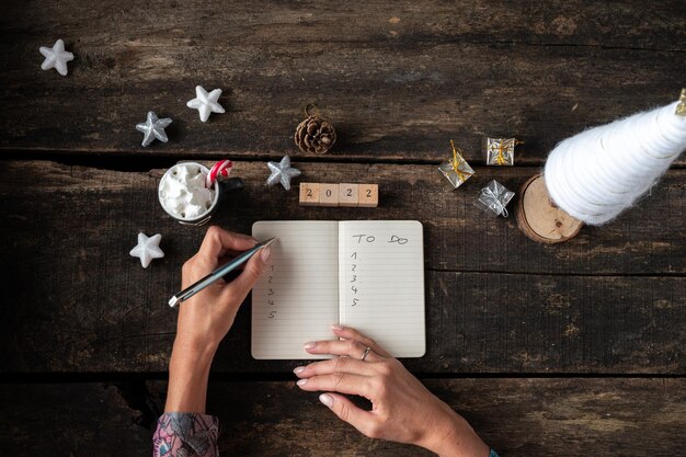 Foto vista dall'alto delle mani femminili che scrivono una lista di cose da fare per l'anno 2022 in un blocco note su una scrivania in legno rustico decorato con decorazioni natalizie.