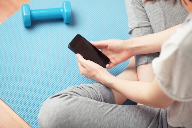 Top view of female person sitting in lotus position and looking at of smartphone screen. Isolation period. Copy space blank display of cellphone. Sports mat and dumbbell. Healthy life style concept.