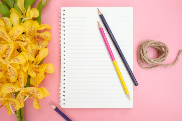 Top view or flat lay of open notebook paper,yellow orchid flowers, color pencils and nature rope on pink background.