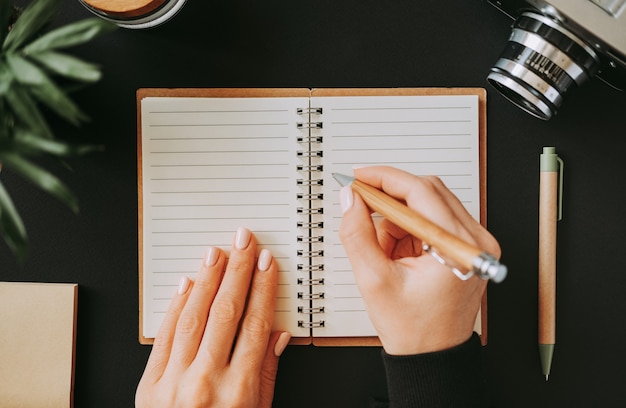 Top view of flat lay woman's hand writes down addresses in an open notebook lying on black table next to camera with pen and notepad