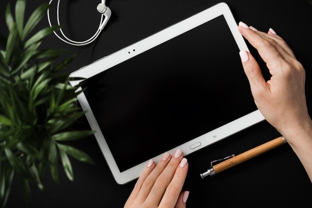 Top view of a flat lay of a womans hands holding a tablet with a black screen lying on a black tabletop next to a pen and headphones. Concept workspace of traveler and blogger. Advertising space