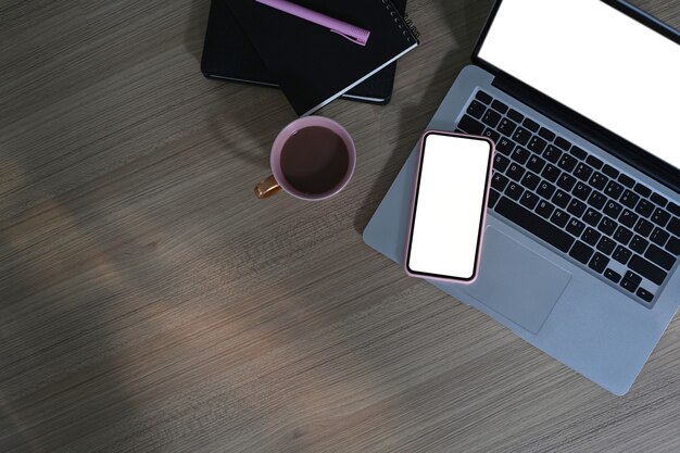 Top view of freelancer workspace with blank screen laptop, mobile phone, coffee cup and notebook on wooden table.