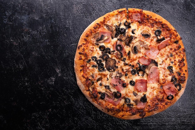 Top view of fresh baked pizza with ham, olives and mushrooms on dark wooden background with copy space available