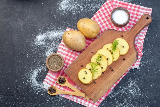 Top view of fresh chopped potatoes with dill pepper on wooden cutting board on red stripped towel spices on dark color background