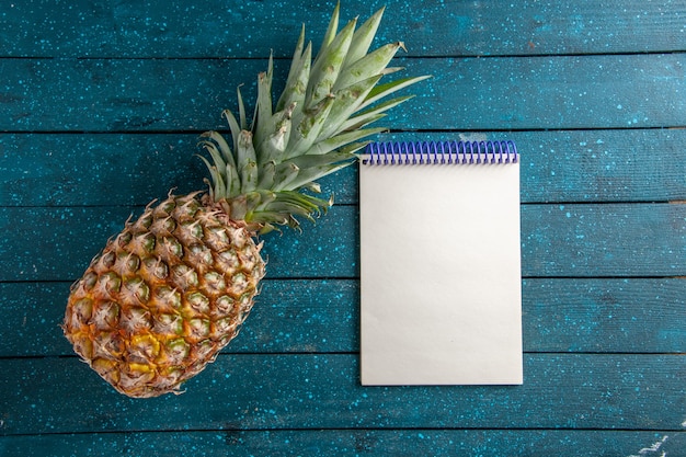 Foto vista dall'alto ananas fresco e blocco note su sfondo di legno blu