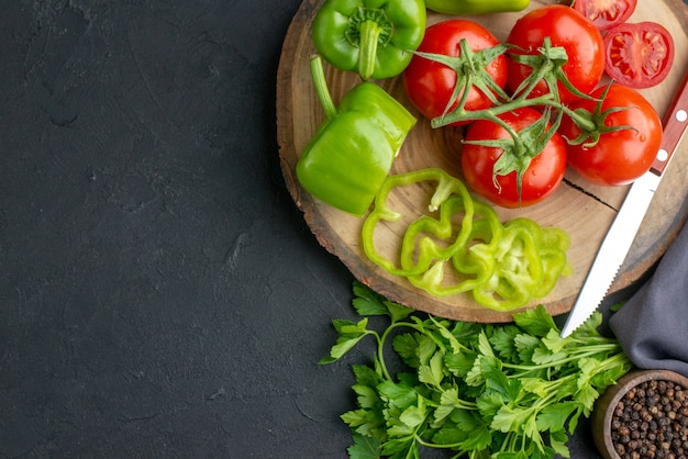 Top view of fresh tomatoes and green peppers on wooden board on the left side on black surface