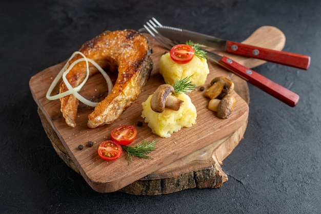 Top view of fried fish meal with mushrooms vegetables cheese and cutlery set on wooden board on black distressed surface