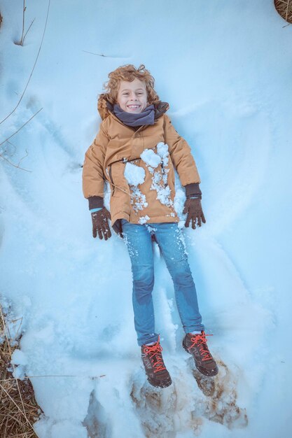 冬の自然の中で楽しみながら、帽子をかぶらずに雪の上に横たわり、天使を作る上着を着た遊び心のある少年の上面図の全身