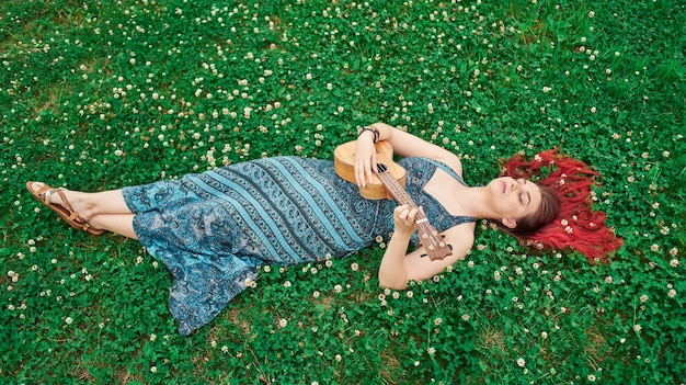 Top view of a full-length woman playing ukulele lying on a meadow in summer, red hair scattered on the grass, eyes closed.