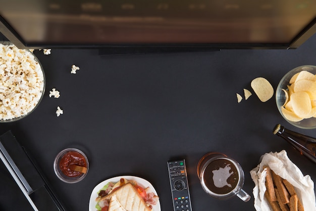 Top view of gamer accessorises and snacks frame, flat lay on black background with copyspace. Joystick and gamepad, keyboard, game console, mouse, mobile phone, beer, chips and popcorn.