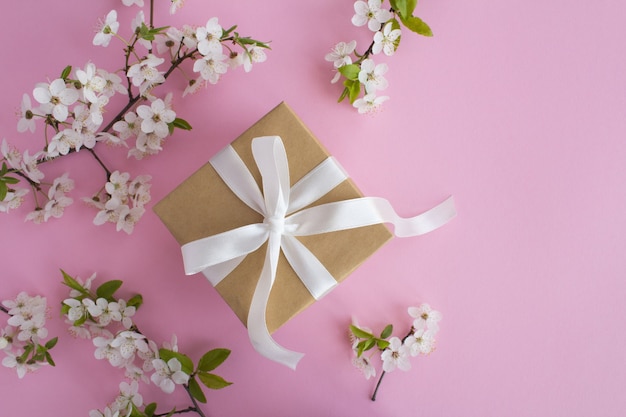 Top view of gift with white bow and flowering tree branches on the pink background