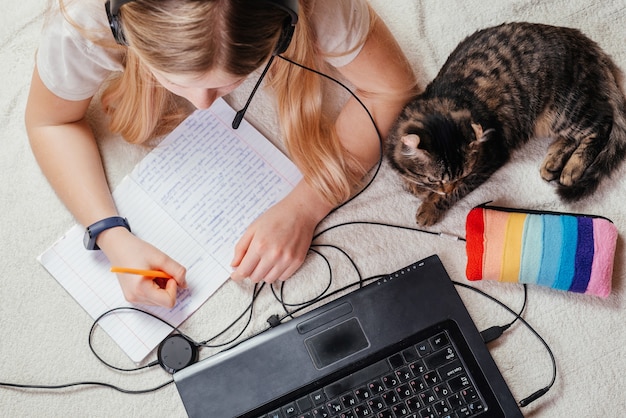 Top view of a girl in headphones using her laptop and making notes in her notebook with a cat by her side
