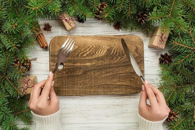 Top view girl holds fork and knife in hand and is ready to eat. Empty wood rectangular plate on wooden board. holiday dinner dish concept with christmas decor