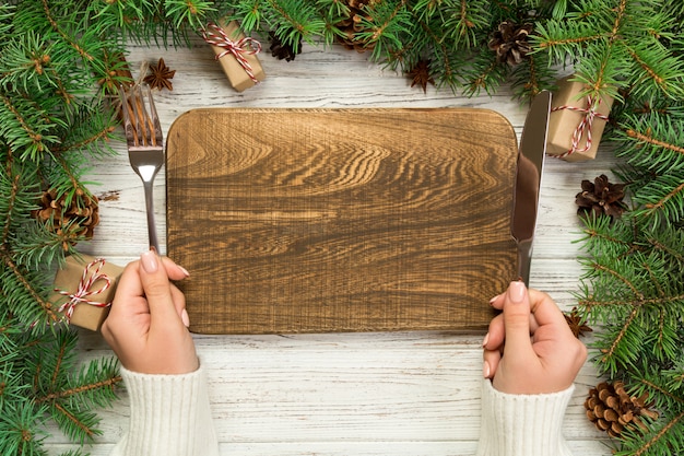 Top view girl holds fork and knife in hand and is ready to eat. Empty wood rectangular plate on wooden christmas . holiday dinner dish  with new year decor