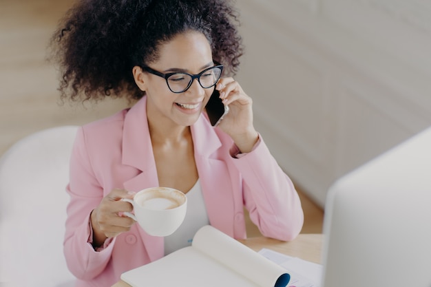 Photo top view of glad dark skinned woman with curly dark hair, has phone conversation, holds mug of drink