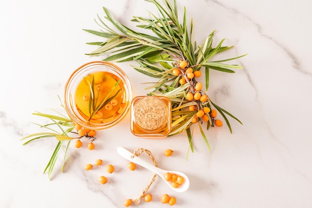 Top view of a glass bottle with a lid and a bowl filled with organic sea buckthorn oil twigs and berries on a marble background a vitamins