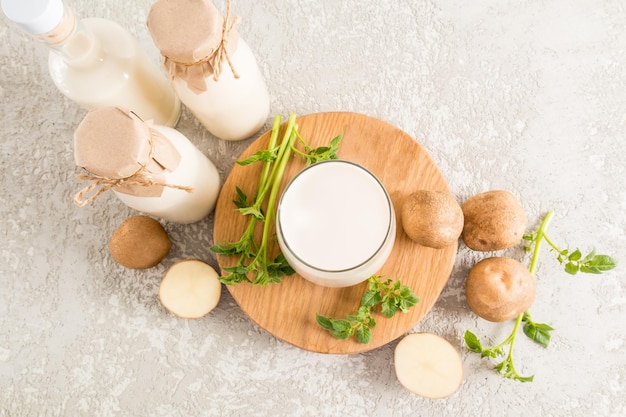 Top view of a glass filled with organic potato milk on a round wooden board two bottles of milk tubers and leaves on a concrete table