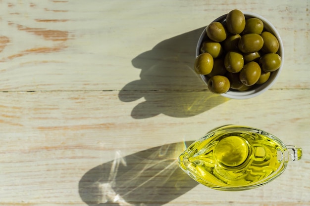 Top view of glass sauceboat with extra virgin olive oil and fresh green olives on wooden table