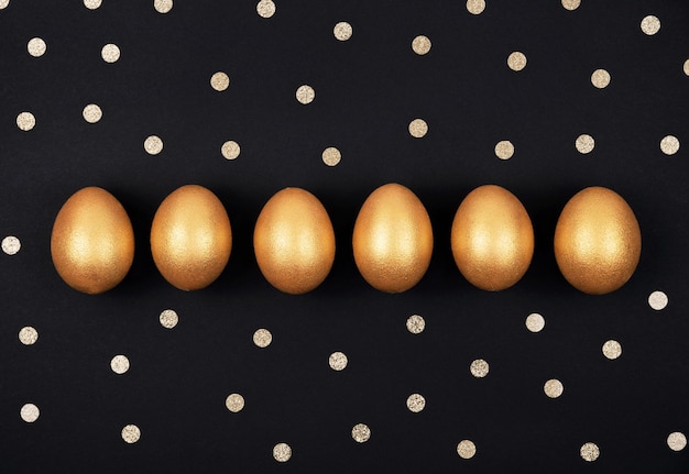 Top view of golden colored easter eggs in a row and confetti on dark black background