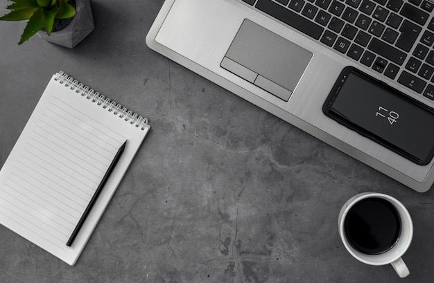 Top view grayscale shot of a laptop, mobile phone, coffee, and notebook on a table