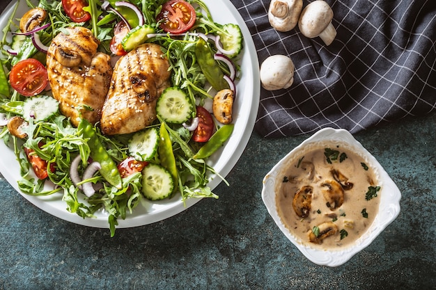 Top view of grilled chicken and mushrooms with salad, cucumber, cherry tomatoes, red onion, with a creamy mushroom sauce in a separate bowl on dark background.