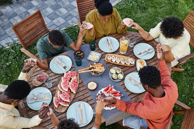 屋外のピクニック テーブルで恵みを言い、手を繋いでいるアフリカ系アメリカ人のグループのトップ ビュー