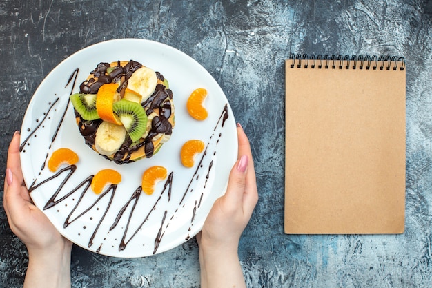 Top view of hand holding American-style pancakes made with natural yogurt and stacked with layers of fruit decorated with chocolate on white plate and notebook