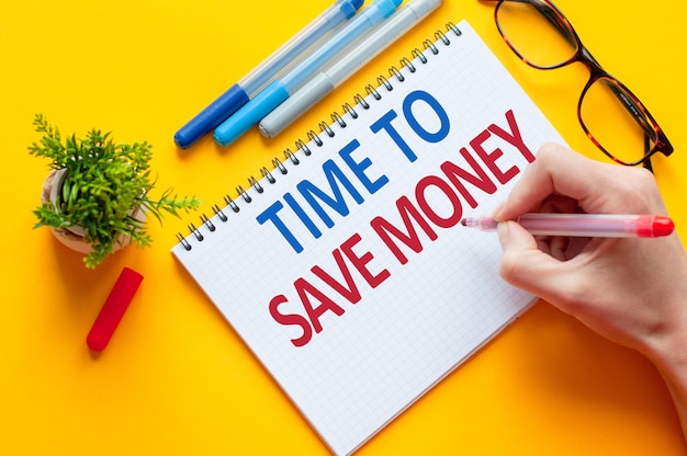 Top view, hand holding pencil writing TIME TO SAVE MONEY list with notebook, pen, glasses, calculator and green flower on yellow table. business and education concept