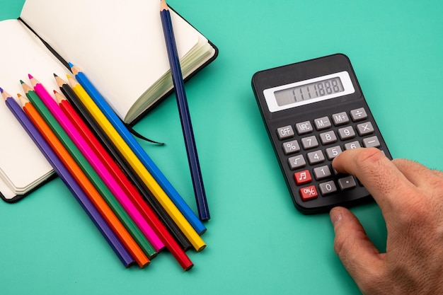 Вид сверху руки, нажимающей кнопки калькулятора на зеленом столе с блокнотом и цветной ручкой