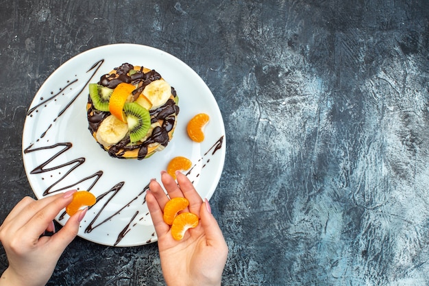 Top view of hand putting tangerines on American-style pancakes made with natural yogurt and stacked with layers of fruit decorated with chocolate on white plate