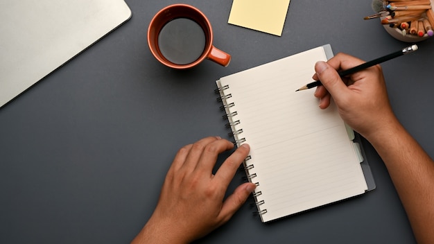 Top view of hand writing on blank notebook on workspace with coffee cup and stationery