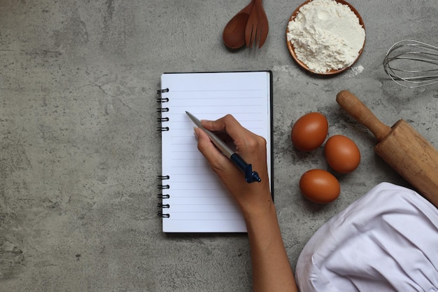 Top view of hand writing on a blank recipe notes with chef hat and cooking ingredients on grey rusti