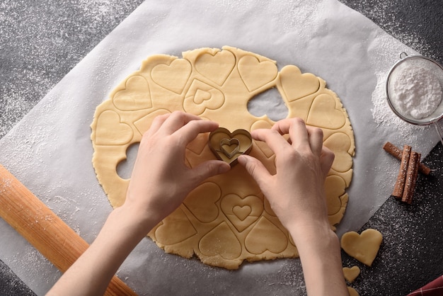 Top view hands cut out hearts from cookie dough on kitchen gray table, cinnamon sticks, rolling pin and powdered sugar