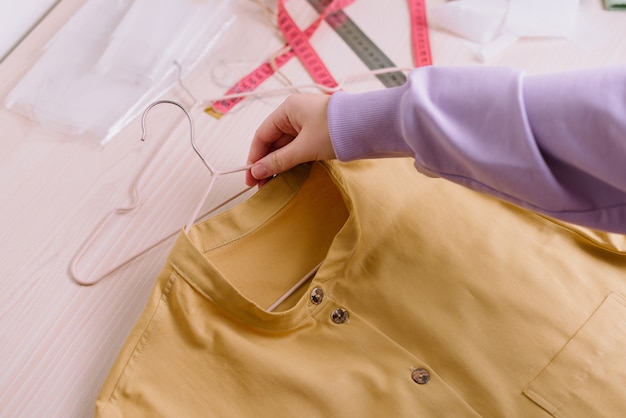 Top view of hands of female seamstress holding finished yellow shirt on a hanger
