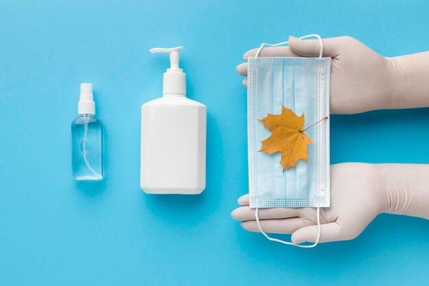사진 뷰 손은 가을 잎 액체 비누 병과 함께 의료 마스크를 들고 있습니다.