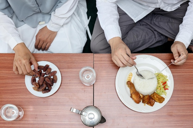 Photo top view of hands takes date fruits or kurma in the white plate and eating rice. ramadan iftar food.